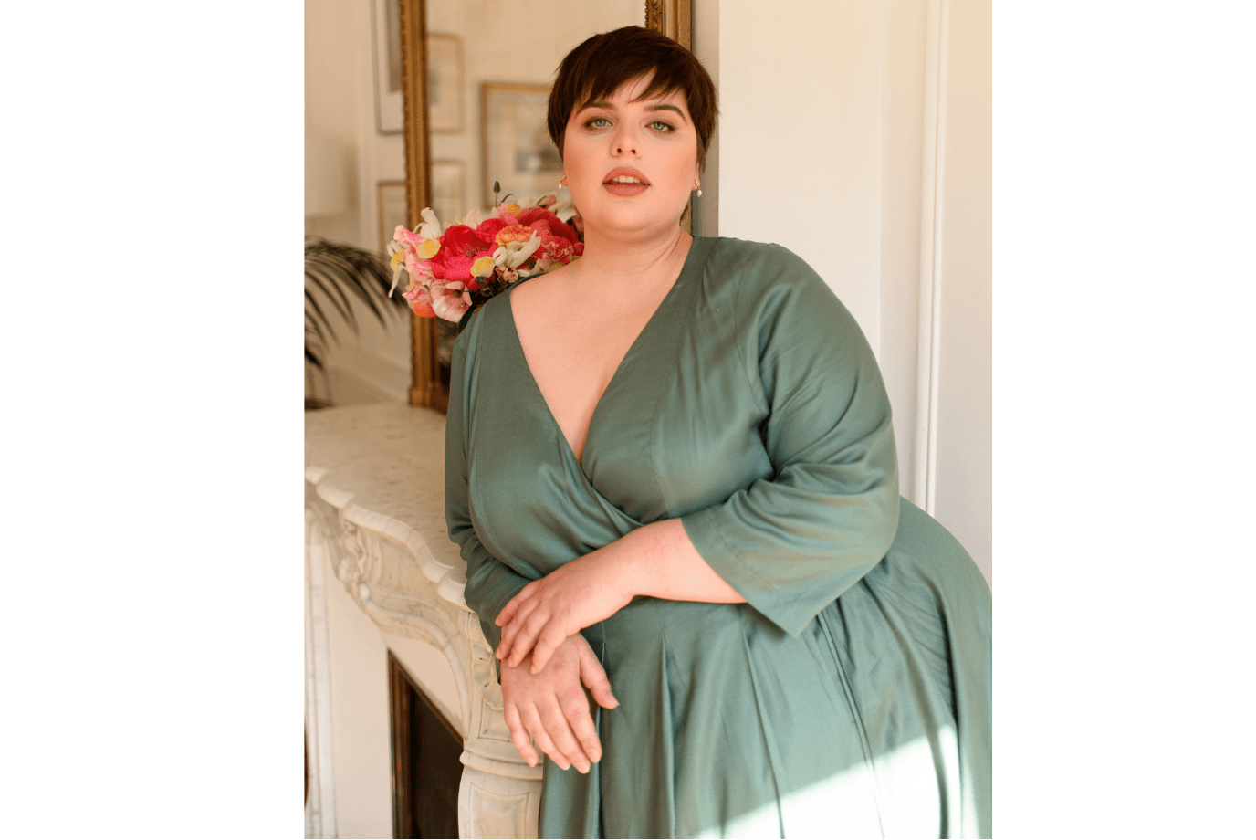Woman wearing a green plus-size wrap dress is leaning against a mantelpiece in a Parisian apartment